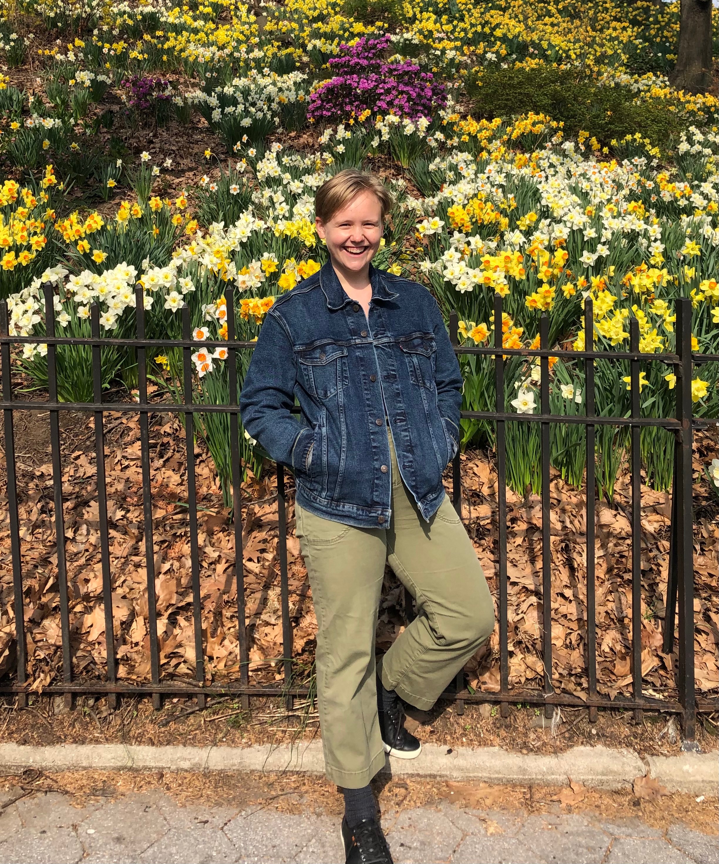 Iz wearing a Jean jacket and army green pants in front of an iron gate with a garden behind it