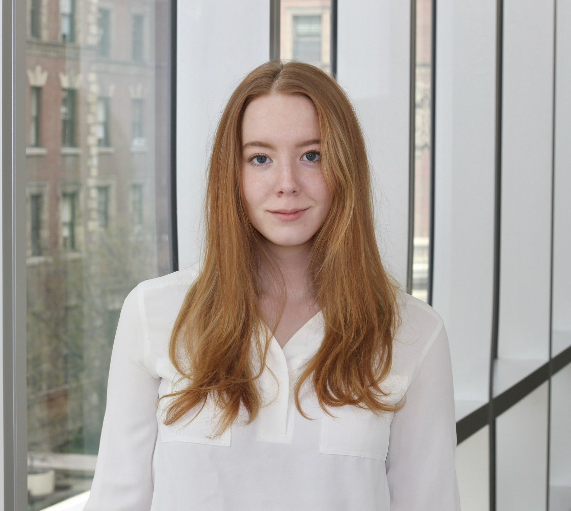 Cassandra wearing a white shirt, standing in front of a window with a brick building outside