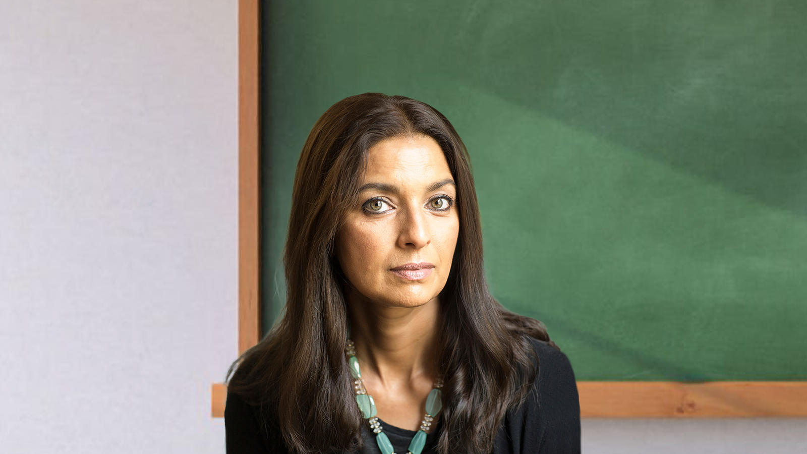 Photo of Bengali American woman, Jhumpa Lahiri, in a dark sweater and turquoise necklace in front of a chalkboard